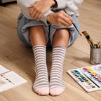 A vertical crop of knees and feet on a wooden floor with watercolor supplies, Sailor Stripe Bamboo in Light Gray Heather socks neatly cuffed at the ankle. [lf]