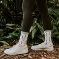 Model in ivory sneakers and crew socks with embroidered lavender sprigs stands on woodchips before tropical foliage, showcasing the Lovely Lavender Bamboo socks in Green Heather. [lf]