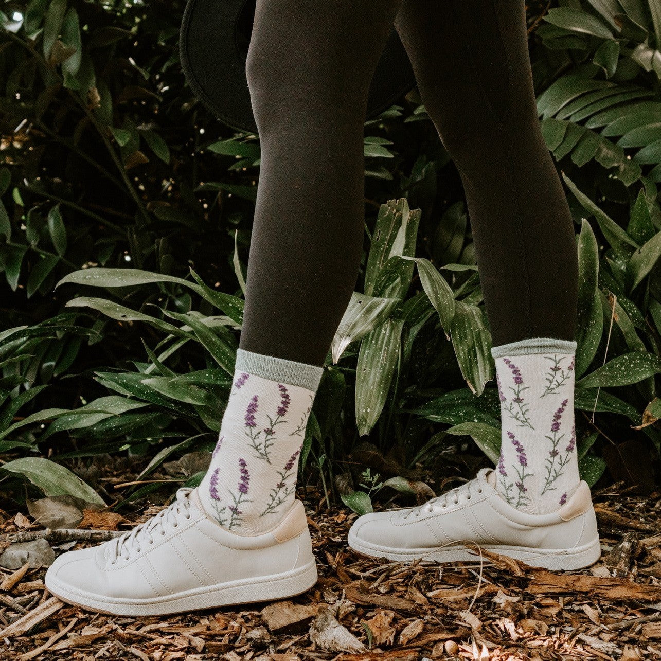 Model in ivory sneakers and crew socks with embroidered lavender sprigs stands on woodchips before tropical foliage, showcasing the Lovely Lavender Bamboo socks in Green Heather. [lf]
