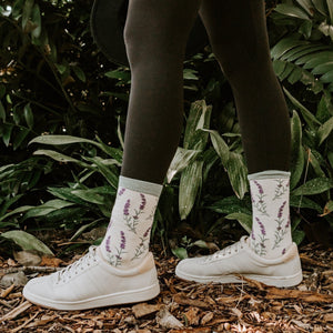 Model in ivory sneakers and crew socks with embroidered lavender sprigs stands on woodchips before tropical foliage, showcasing the Lovely Lavender Bamboo socks in Green Heather. [lf]