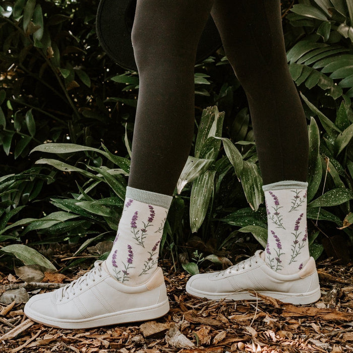 Model in ivory sneakers and crew socks with embroidered lavender sprigs stands on woodchips before tropical foliage, showcasing the Lovely Lavender Bamboo socks in Green Heather. [lf]