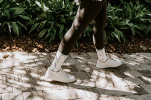 Mid-stride on a dappled brick path, ivory sneakers and lavender-print socks peek beneath dark leggings, featuring the Lovely Lavender Bamboo socks in Green Heather. [lf]