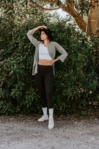 Woman in olive jacket, cropped tee, leggings and tall ivory boot-socks poses by a hedge, styled with the Lovely Lavender Bamboo socks in Green Heather. [lf]