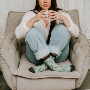 Calm morning portrait beside pampas grass and monstera, hands wrapped around a white mug, feet in mint lavender socks resting on the cushion in the Lovely Lavender Bamboo socks in Green Heather. [lf]