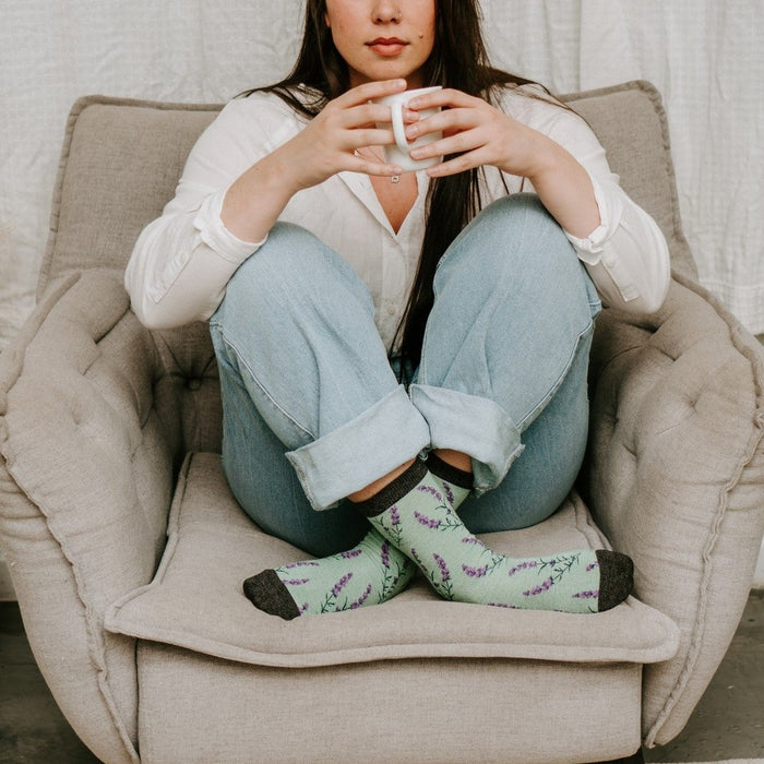 Calm morning portrait beside pampas grass and monstera, hands wrapped around a white mug, feet in mint lavender socks resting on the cushion in the Lovely Lavender Bamboo socks in Green Heather. [lf]