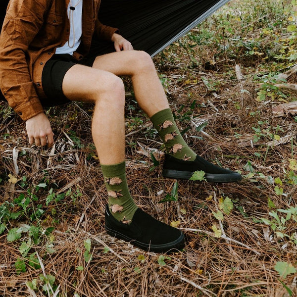 Man in a wide-brim hat and suede jacket sits smiling on a hammock, wearing Armadillos socks in Green. [lf]