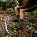 Man in a brown suede jacket leans forward on the edge of a hammock, smiling, wearing Armadillos socks in Green. [lf]
