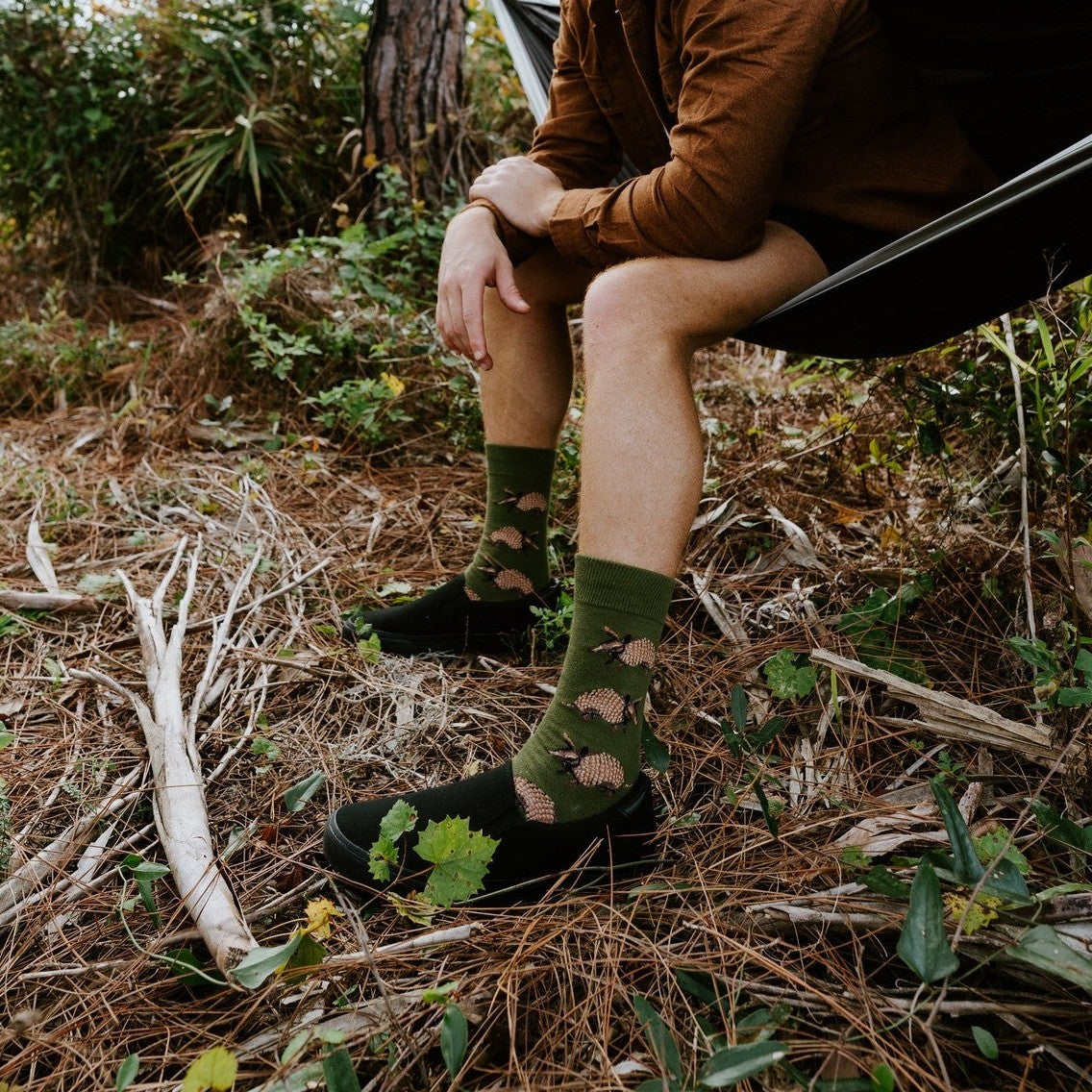 Man in a brown suede jacket leans forward on the edge of a hammock, smiling, wearing Armadillos socks in Green. [lf]