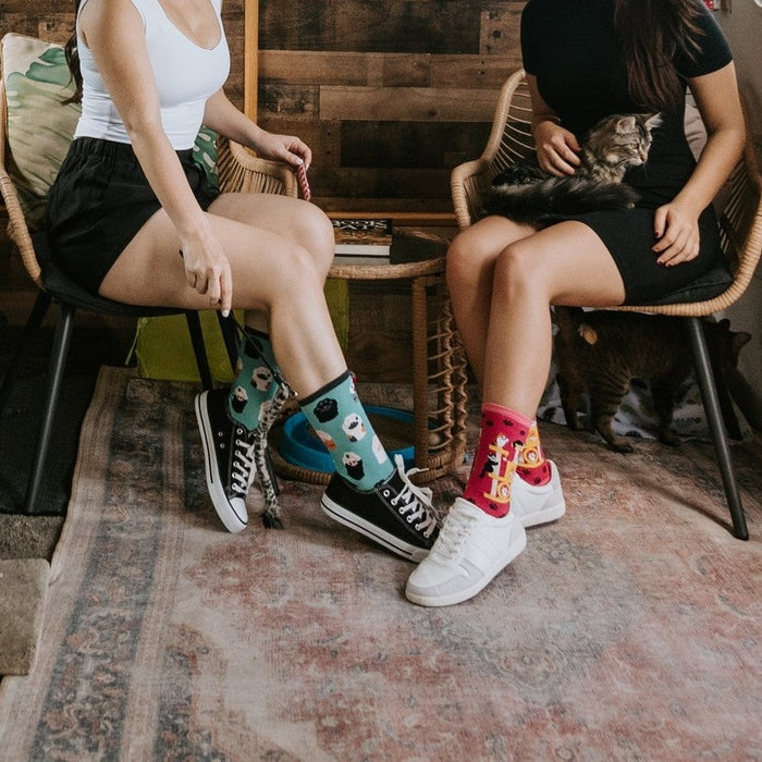 Two friends sit in matching rattan chairs in the camper lounge while a black cat prowls the counter and a fluffy tabby is held in a lap, one wearing Toe Beans high tops and the other wearing Cool Cats Club socks in Pink. [lf]