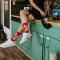 Woman sits on a green kitchen counter petting a long-haired tabby cat, a striped kitten on the floor nearby, while wearing Cool Cats Club socks in Pink. [lf]
