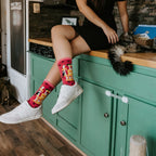 Woman sits on a green kitchen counter petting a long-haired tabby cat, a striped kitten on the floor nearby, while wearing Cool Cats Club socks in Pink. [lf]