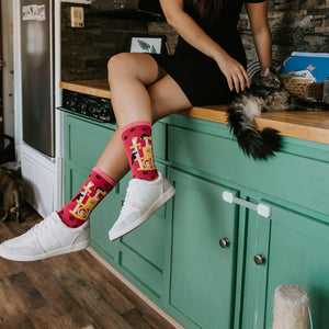 Woman sits on a green kitchen counter petting a long-haired tabby cat, a striped kitten on the floor nearby, while wearing Cool Cats Club socks in Pink. [lf]