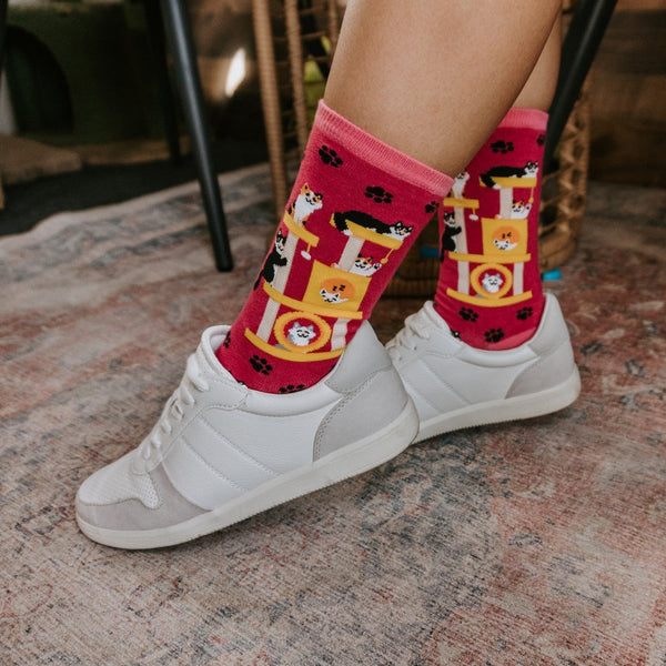 Close-up of crossed ankles in white sneakers on a faded oriental rug, focusing on the cat-tower and paw-print design of Cool Cats Club socks in Pink. [lf]