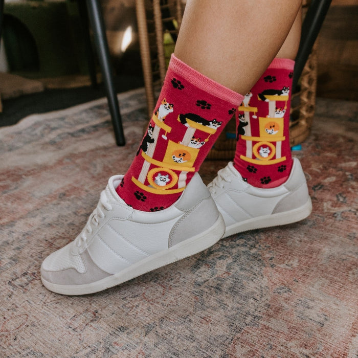 Close-up of crossed ankles in white sneakers on a faded oriental rug, focusing on the cat-tower and paw-print design of Cool Cats Club socks in Pink. [lf]
