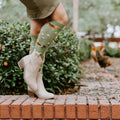 Close-up of cream cowboy boots and Chicken Ranch socks featuring tiny printed roosters and hens, with real chickens pecking on the path behind. [lf]