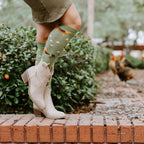 Close-up of cream cowboy boots and Chicken Ranch socks featuring tiny printed roosters and hens, with real chickens pecking on the path behind. [lf]