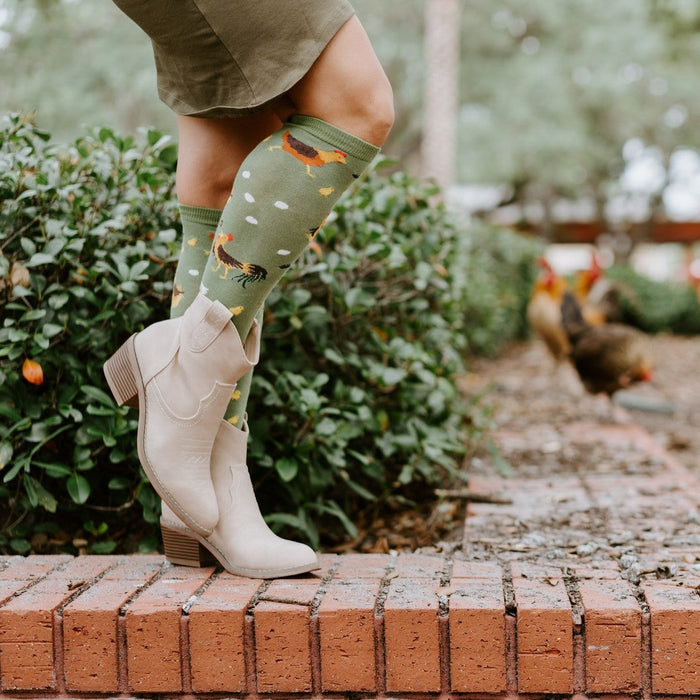 Close-up of cream cowboy boots and Chicken Ranch socks featuring tiny printed roosters and hens, with real chickens pecking on the path behind. [lf]