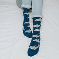 A close-in view of a smiling reader with her legs outstretched on a white duvet, the dolphin-print Joyous Dolphin Bamboo in Blue Heather socks catching the light. [lf]