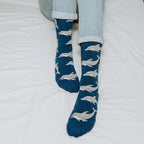 A close-in view of a smiling reader with her legs outstretched on a white duvet, the dolphin-print Joyous Dolphin Bamboo in Blue Heather socks catching the light. [lf]
