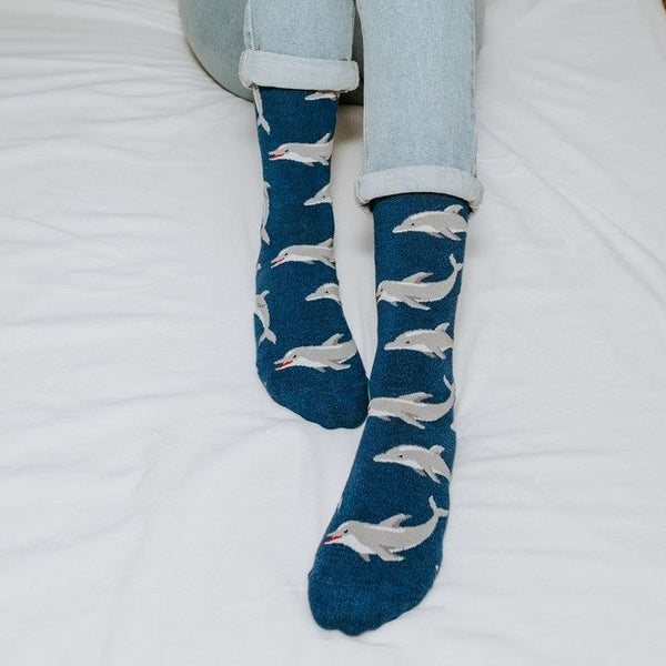 A close-in view of a smiling reader with her legs outstretched on a white duvet, the dolphin-print Joyous Dolphin Bamboo in Blue Heather socks catching the light. [lf]