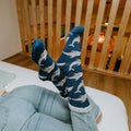 An overhead shot of a woman lying with her legs raised against a slatted wood divider, Joyous Dolphin Bamboo in Blue Heather socks crossed at the ankle. [lf]