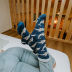 An overhead shot of a woman lying with her legs raised against a slatted wood divider, Joyous Dolphin Bamboo in Blue Heather socks crossed at the ankle. [lf]