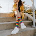 A woman with curly hair in a yellow button-down sits on concrete steps, her Pride Stripes rainbow crew socks paired with white sneakers as she rests her chin on her hand. [lf]