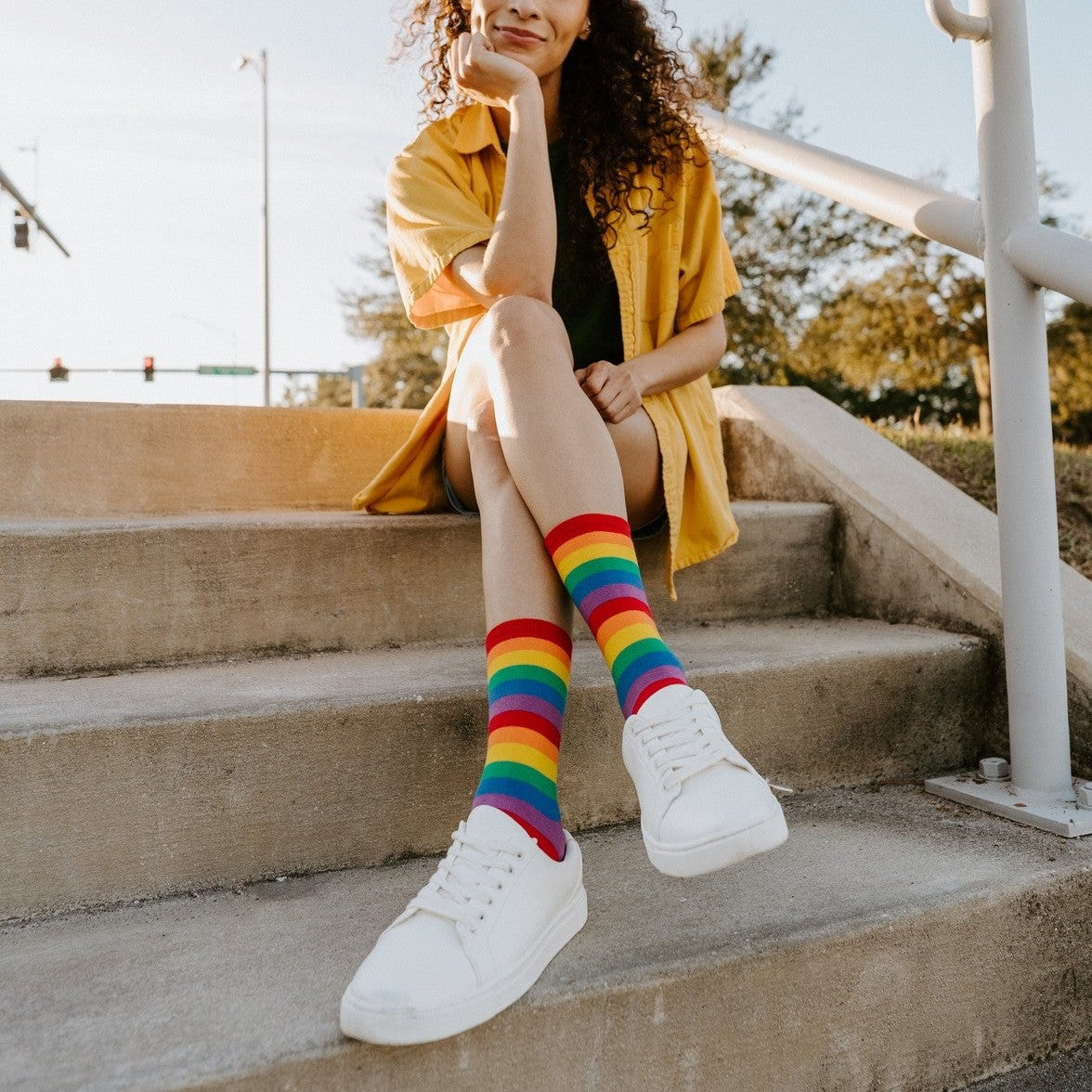 A woman with curly hair in a yellow button-down sits on concrete steps, her Pride Stripes rainbow crew socks paired with white sneakers as she rests her chin on her hand. [lf]