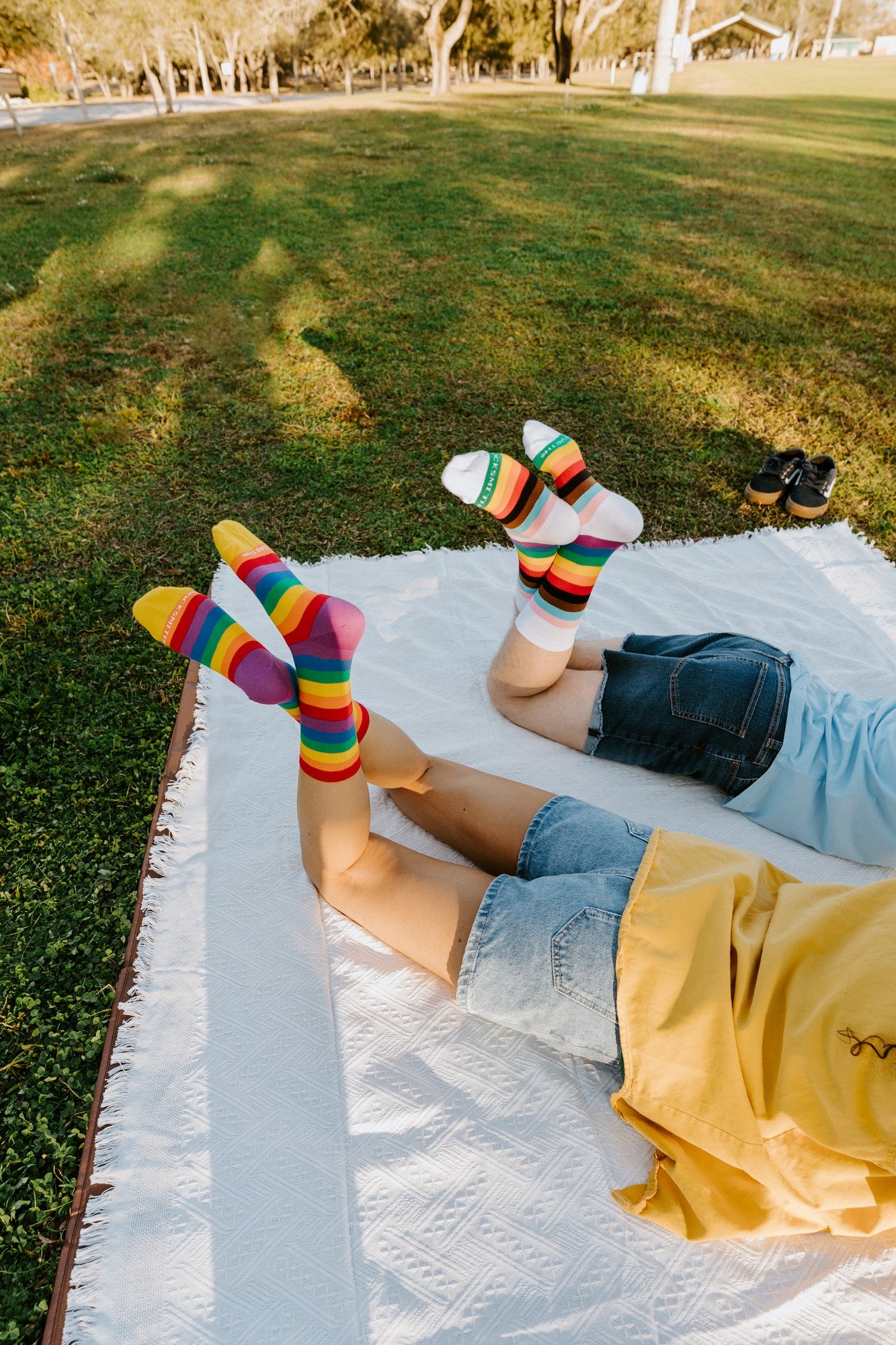 Two people lie back on a white picnic blanket with feet in the air, showing off Progress Stripes socks above and the Pride Stripes rainbow socks alongside. [lf]