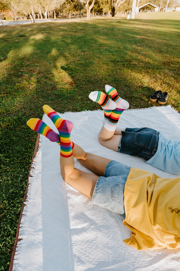 Two people lie back on a white picnic blanket with feet in the air, showing off Progress Stripes socks above and the Pride Stripes rainbow socks alongside. [lf]