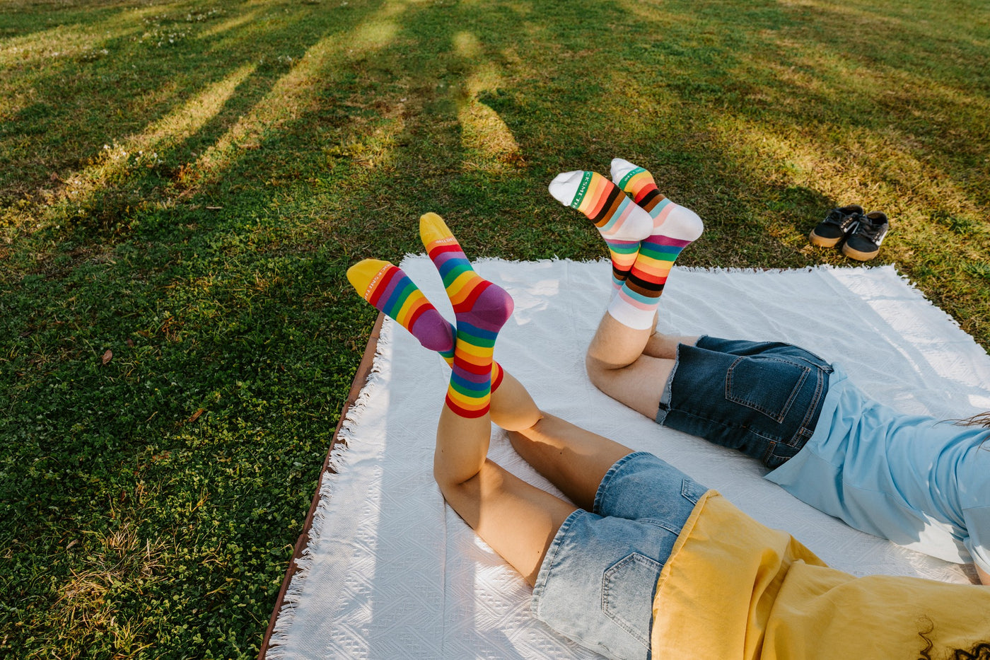 Wider angle of the picnic scene, legs raised on the blanket so the Progress Stripes and Pride Stripes socks catch the afternoon sun. [lf]