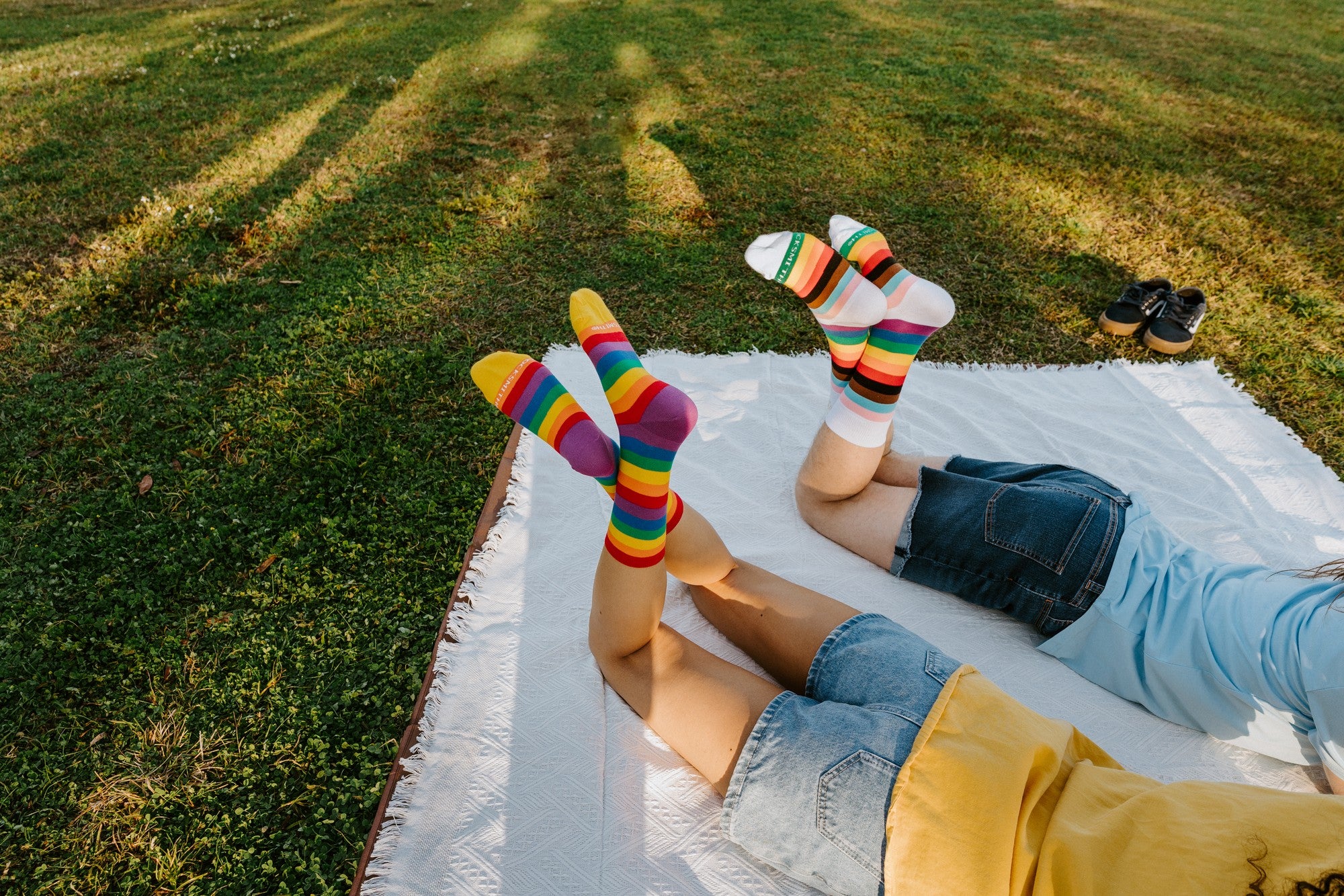 Wider angle of the picnic scene, legs raised on the blanket so the Progress Stripes and Pride Stripes socks catch the afternoon sun. [lf]