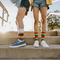 Close-up of two pairs of feet on a concrete step, contrasting the Progress Stripes design with the bold rainbow of the Pride Stripes socks. [lf]