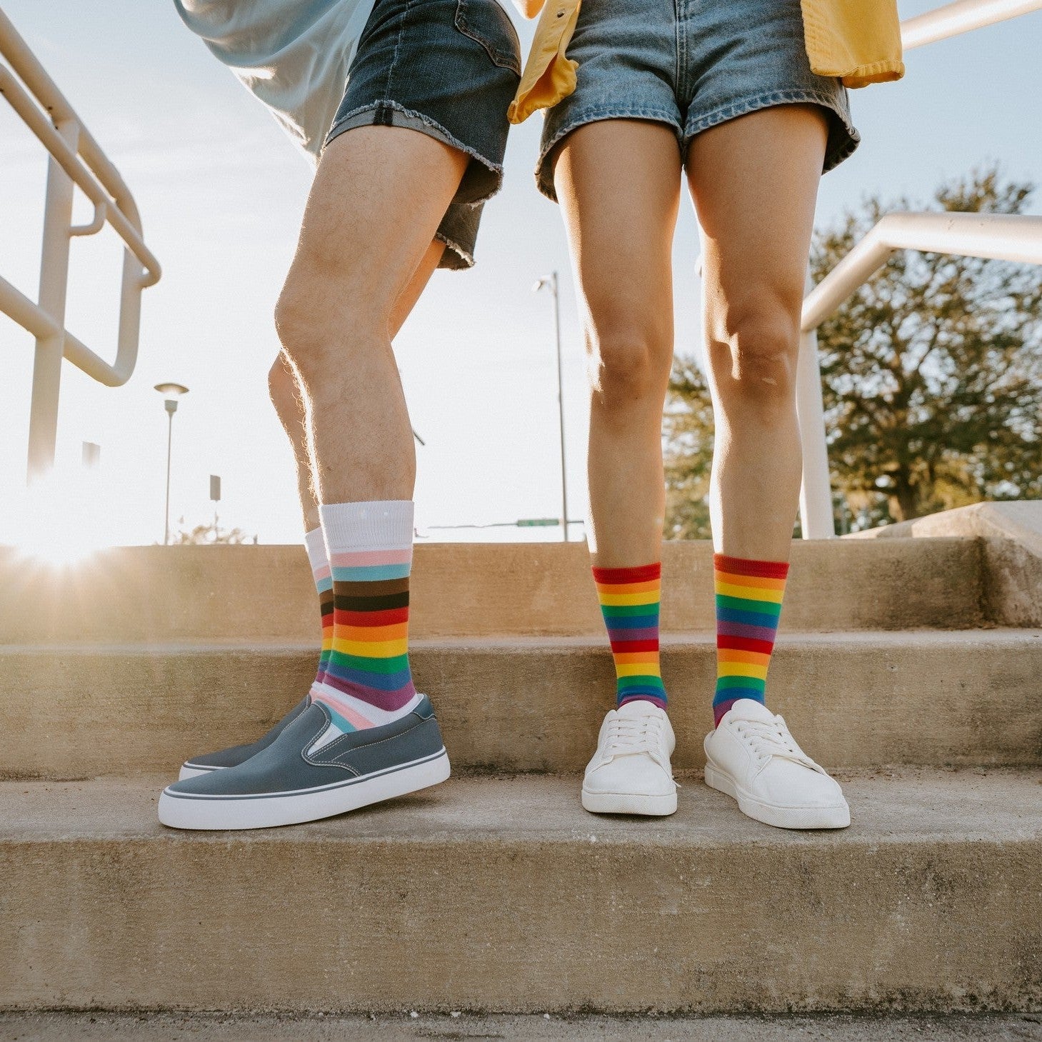Close-up of two pairs of feet on a concrete step, contrasting the Progress Stripes design with the bold rainbow of the Pride Stripes socks. [lf]