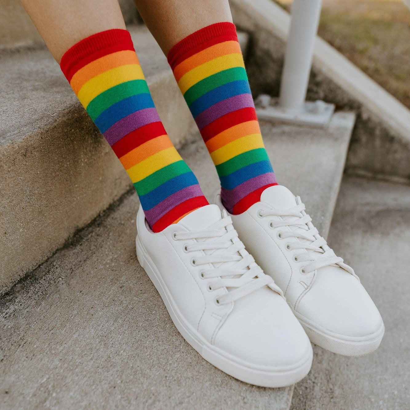 Close-up of crossed legs on concrete steps showing the bright red, orange, yellow, green, blue, and purple bands of the Pride Stripes socks above clean white sneakers. [lf]