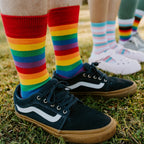 Close-up of three pairs of feet on the grass showing the Pride Stripes rainbow socks beside Trans Stripes and Progress Stripes designs. [lf]