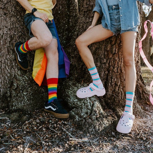Two friends lean against the wide oak trunk side by side, their crossed legs framing Trans Stripes socks in Multi in bold rainbow and pastel pink, white and blue stripes. [lf]