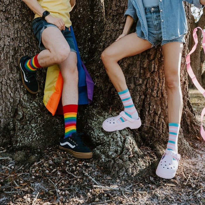 Two friends lean against the wide oak trunk side by side, their crossed legs framing Trans Stripes socks in Multi in bold rainbow and pastel pink, white and blue stripes. [lf]