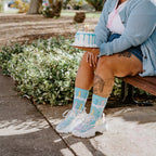 Woman in a light blue cardigan and denim skirt sits on a park bench holding a small white birthday cake with blue drip icing and lit candles, wearing Birthday Candles socks in Blue and iridescent platform sneakers. [lf]