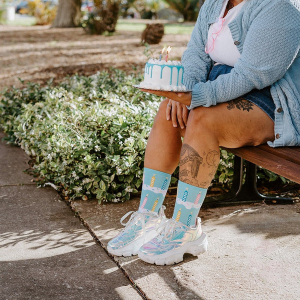 Woman in a light blue cardigan and denim skirt sits on a park bench holding a small white birthday cake with blue drip icing and lit candles, wearing Birthday Candles socks in Blue and iridescent platform sneakers. [lf]