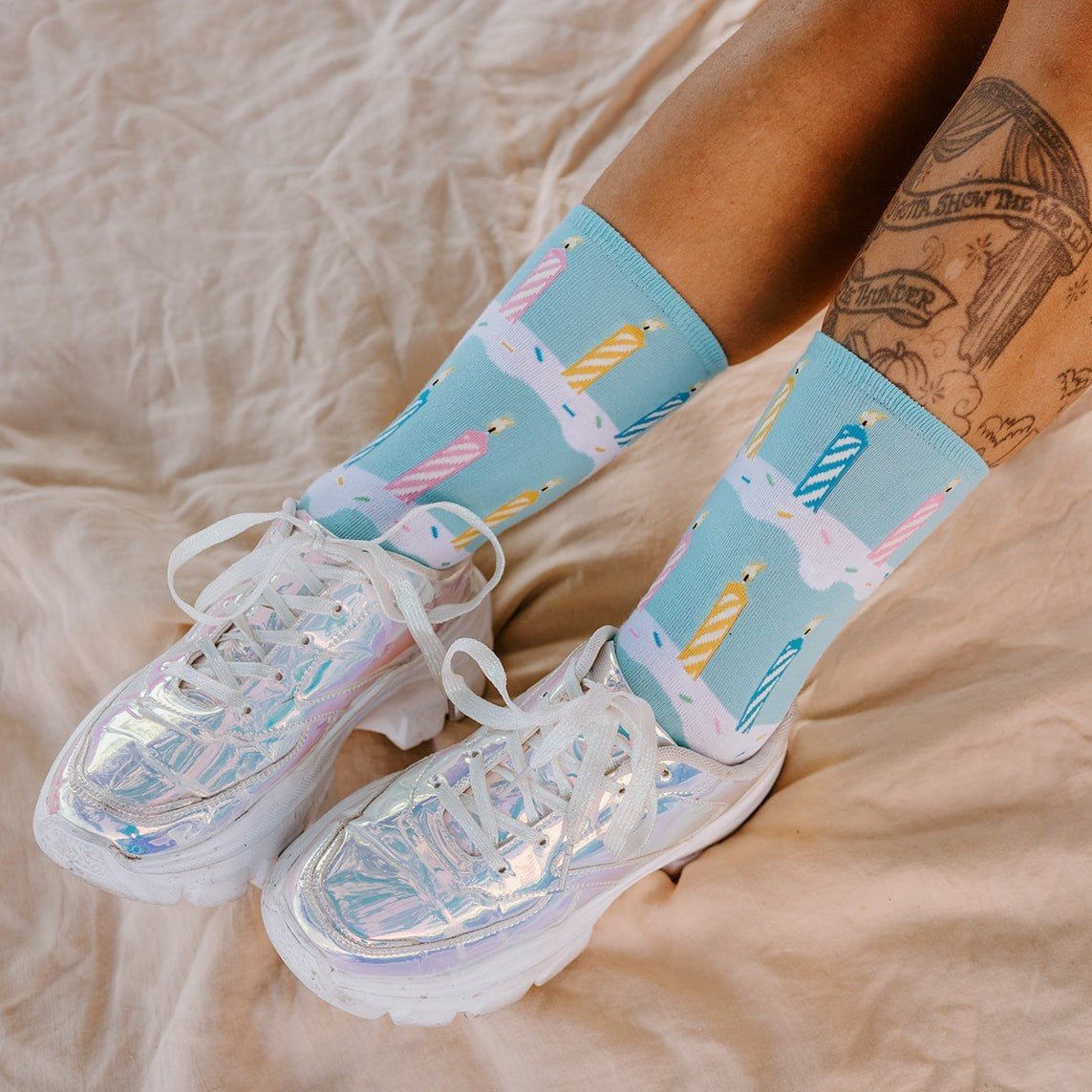 Studio-style flat lay on rumpled cream bedding showcasing crossed legs in Birthday Candles socks in Blue paired with iridescent platform sneakers, candle and drip-icing artwork in full view. [lf]