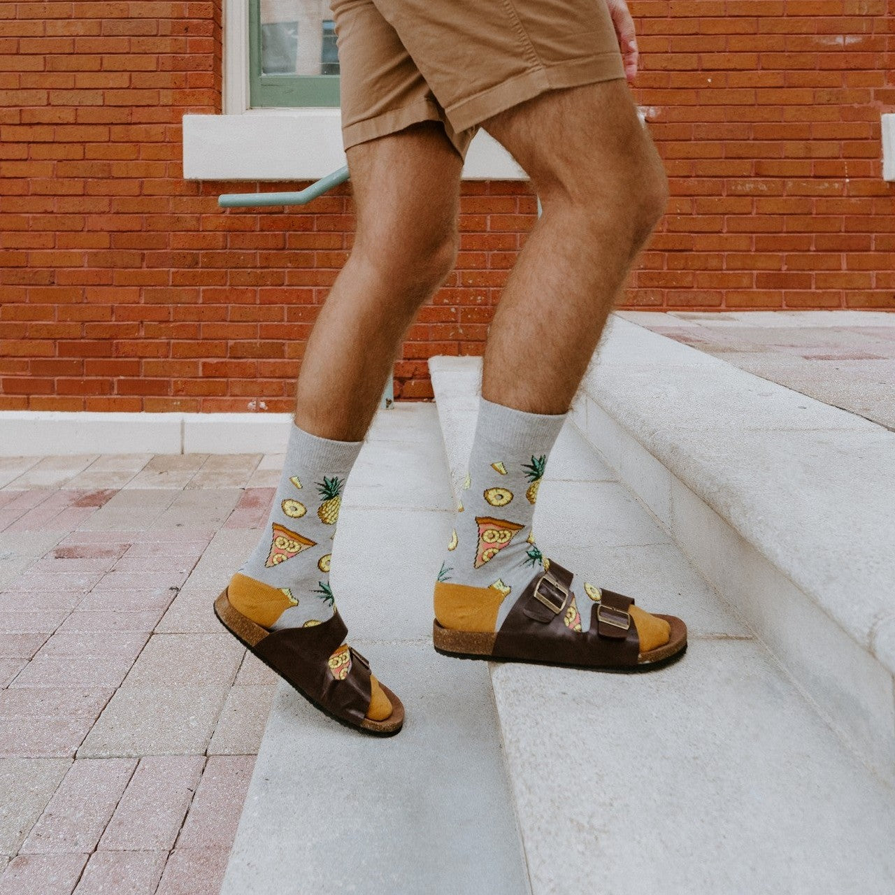 Close-up of a man's legs in tan shorts stepping up white concrete stairs in front of a red brick building, his Hawaiian Pizza socks showing pineapple and pizza prints above brown buckle sandals with mustard footbeds. [lf]