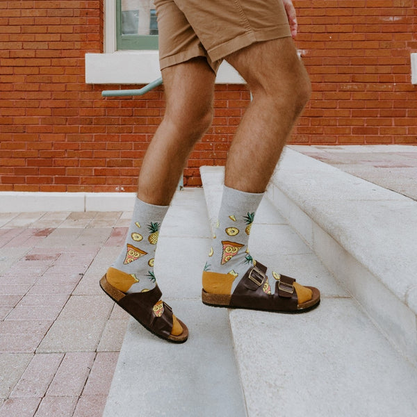 Close-up of a man's legs in tan shorts stepping up white concrete stairs in front of a red brick building, his Hawaiian Pizza socks showing pineapple and pizza prints above brown buckle sandals with mustard footbeds. [lf]
