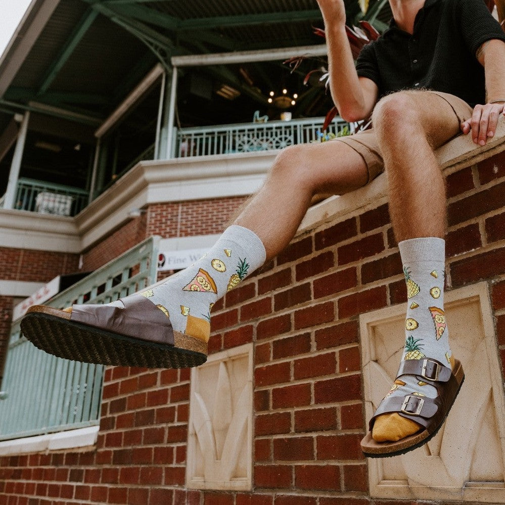 Low angle view of a bearded man in black shirt and tan shorts seated atop a red brick wall holding a pizza slice, palm fronds overhead, his Hawaiian Pizza socks visible above brown sandals. [lf]