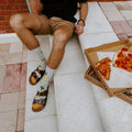 Overhead view of a man in tan shorts seated on white concrete with an open pizza box revealing a half-eaten pepperoni pizza beside him, his Hawaiian Pizza socks with pineapple prints visible above brown buckle sandals on a red brick patio. [lf]