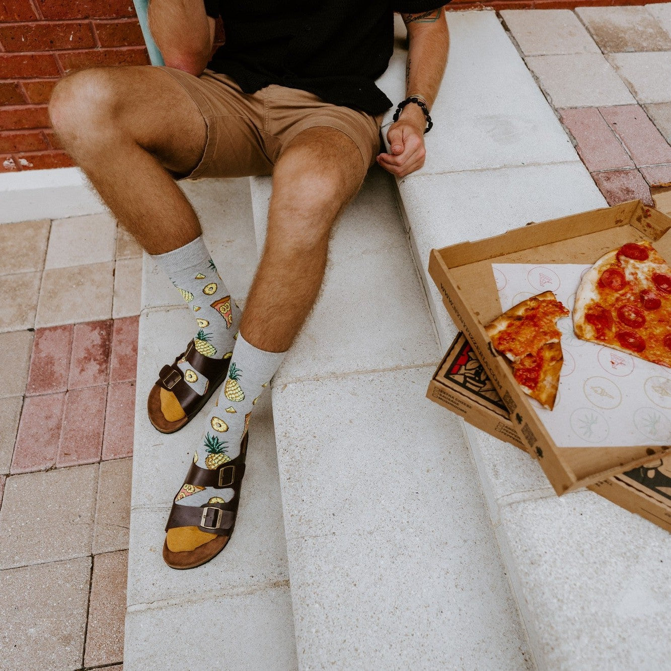 Overhead view of a man in tan shorts seated on white concrete with an open pizza box revealing a half-eaten pepperoni pizza beside him, his Hawaiian Pizza socks with pineapple prints visible above brown buckle sandals on a red brick patio. [lf]