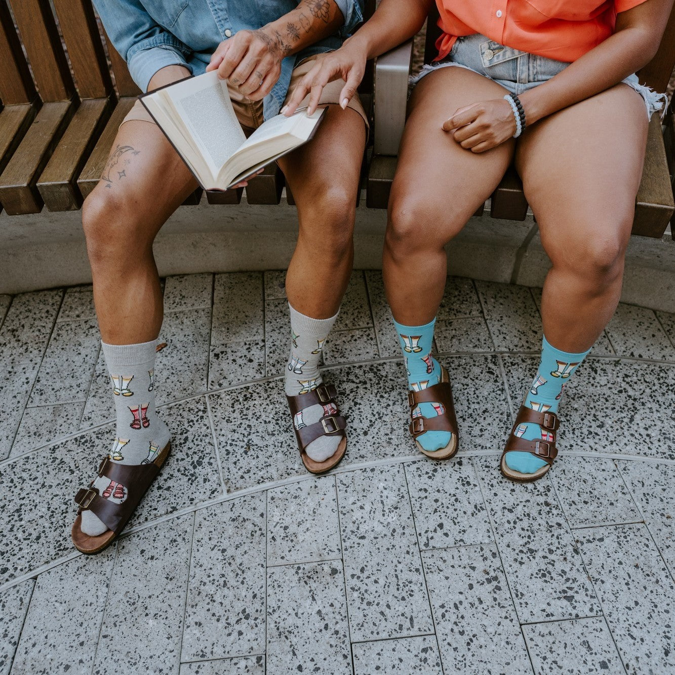 A close crop of four legs side by side on the bench, the cuffed Socks And Sandals in Blue paired with brown leather sandals on a speckled terrazzo floor. [lf]