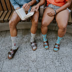 A close crop of four legs side by side on the bench, the cuffed Socks And Sandals in Blue paired with brown leather sandals on a speckled terrazzo floor. [lf]