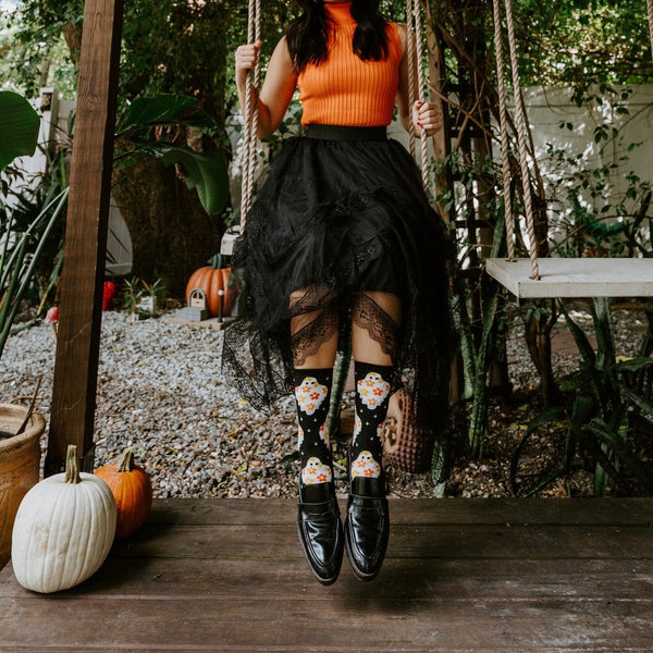 Model on a wooden swing surrounded by greenery and pumpkins, kicking out her feet in Flower Ghosts socks in Black. [lf]