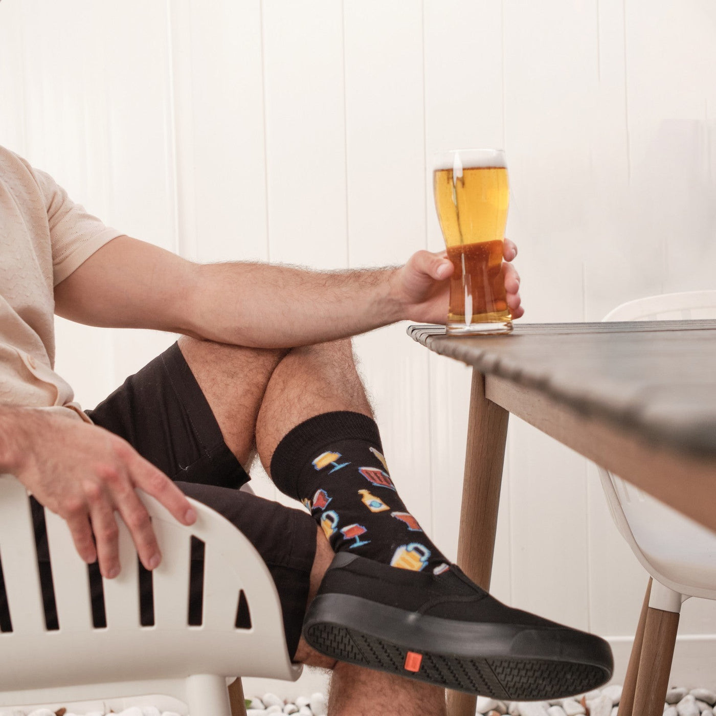 Man in a cream knit polo sits in a slatted patio chair holding a pint of beer toward the table, wearing Get Your Beer Glasses socks in Black with black slip-on sneakers. [lf]